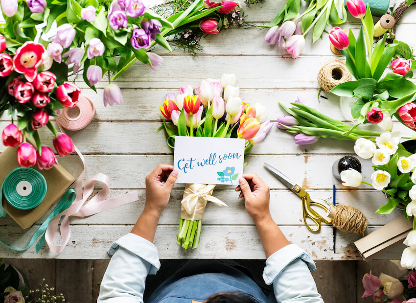 A person holding a bouquet of tulips with a "Get well soon" card, surrounded by colorful flowers and gift-wrapping materials on a wooden table A person holding a bouquet of tulips with a "Get well soon" card, surrounded by colorful flowers and gift-wrapping materials on a wooden table